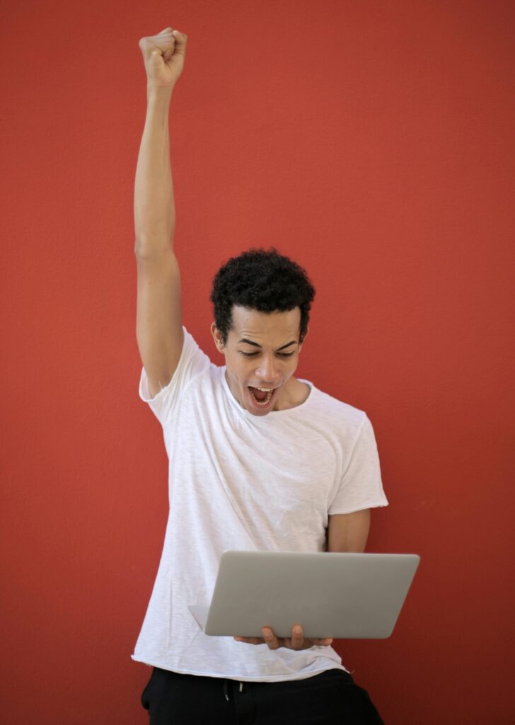 A young man in a white shirt celebrates success holding a laptop against a red background.